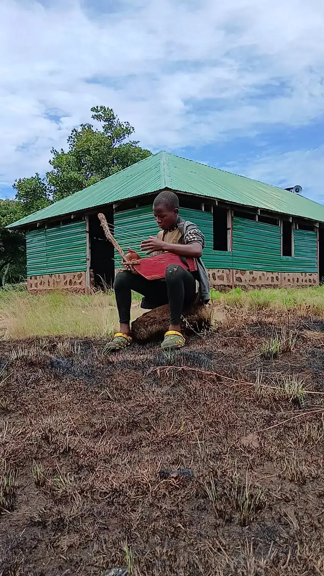 A boy sits outdoors playing a traditional stringed instrument in front of a green-roofed building