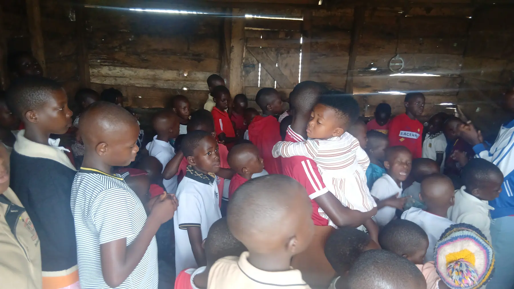 A crowd of children gathered closely together inside a timber hall, some smiling towards the camera