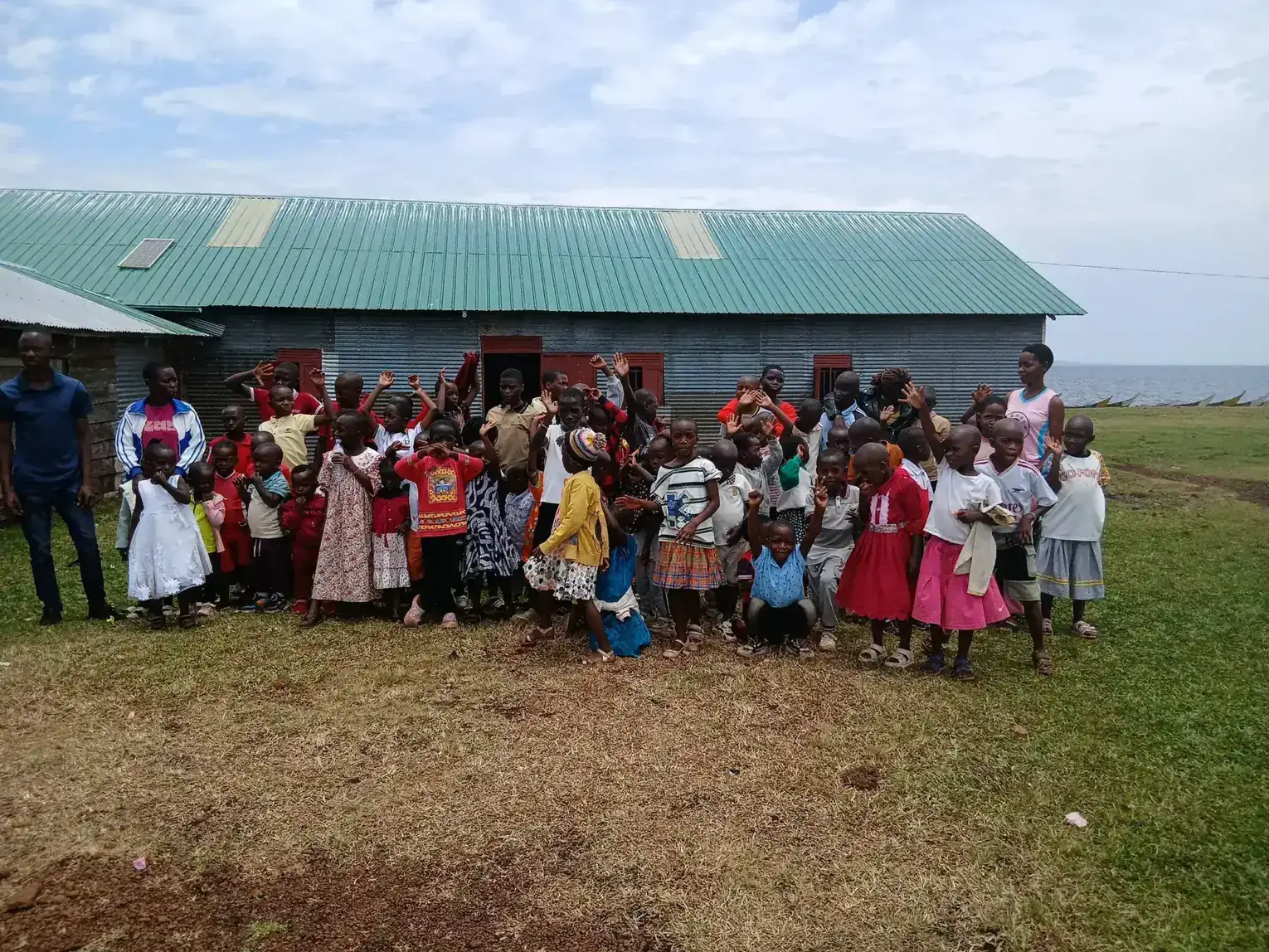 Children and adults stand together outside a green-roofed hall, many waving at the camera