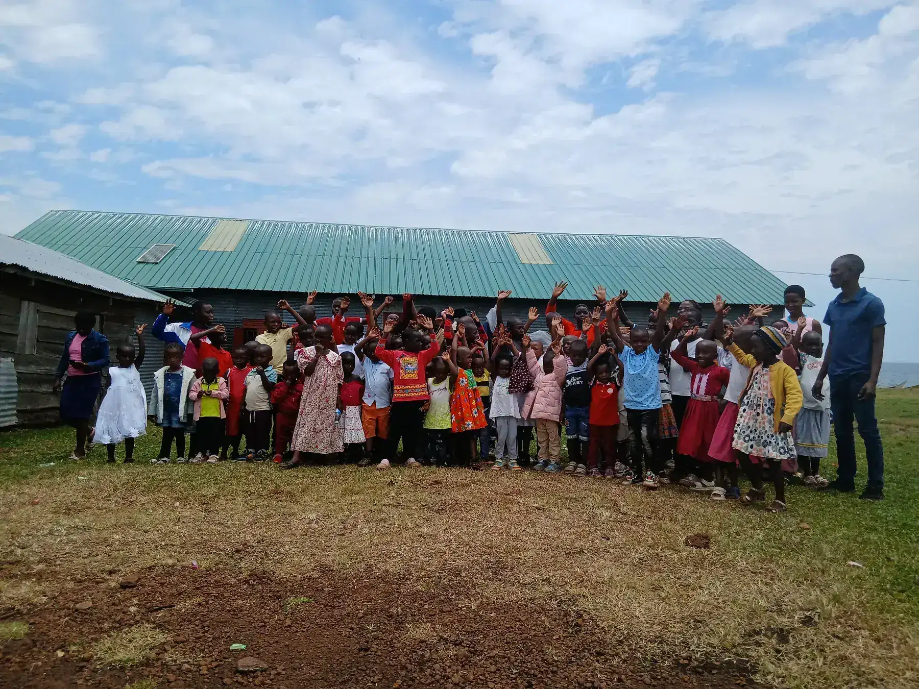 Dozens of children in bright clothes waving alongside adult leaders in front of a green-roofed hall in the Ssese Islands