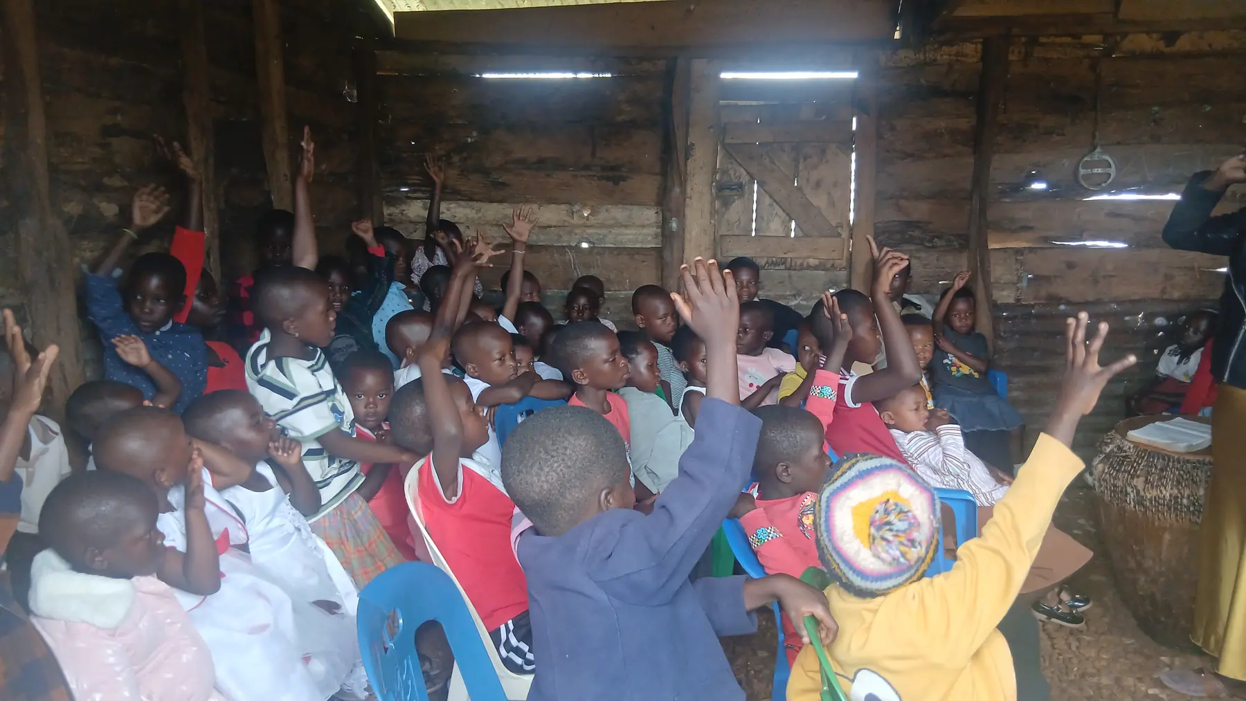Children seated in rows raising their hands during a session inside a timber hall