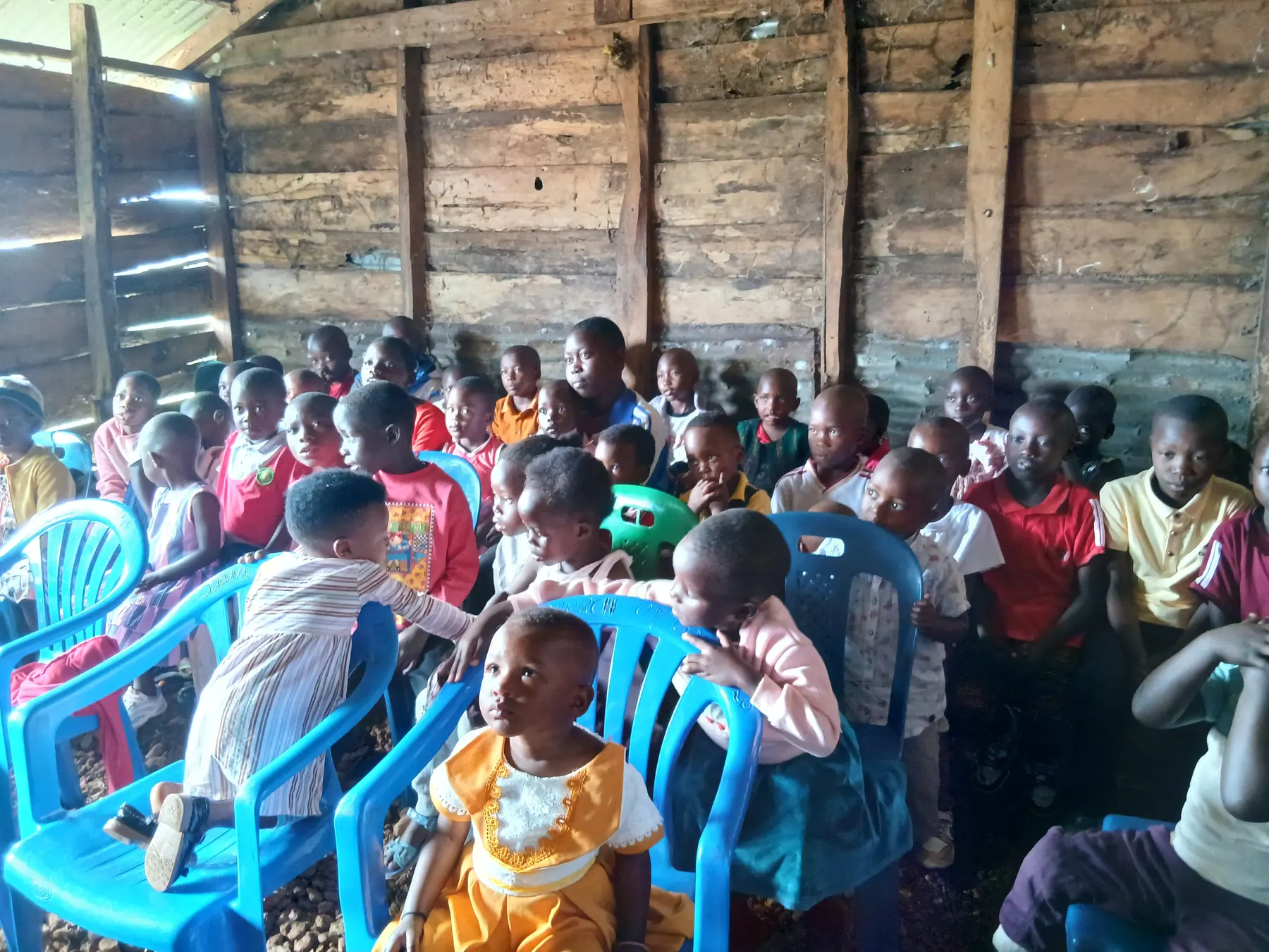 Children sit close together on chairs inside a timber church hall, listening