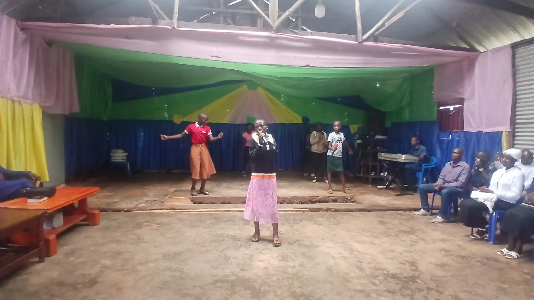 Two girls perform for an audience at the front of a church hall decorated in green, yellow and blue