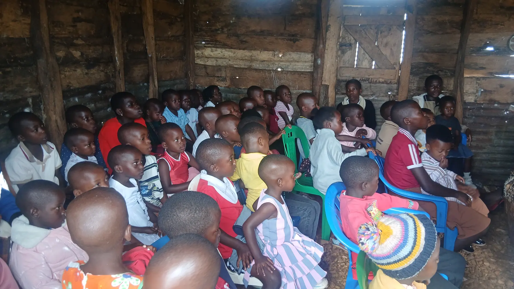 Rows of children seated attentively on blue plastic chairs during a session in a timber hall