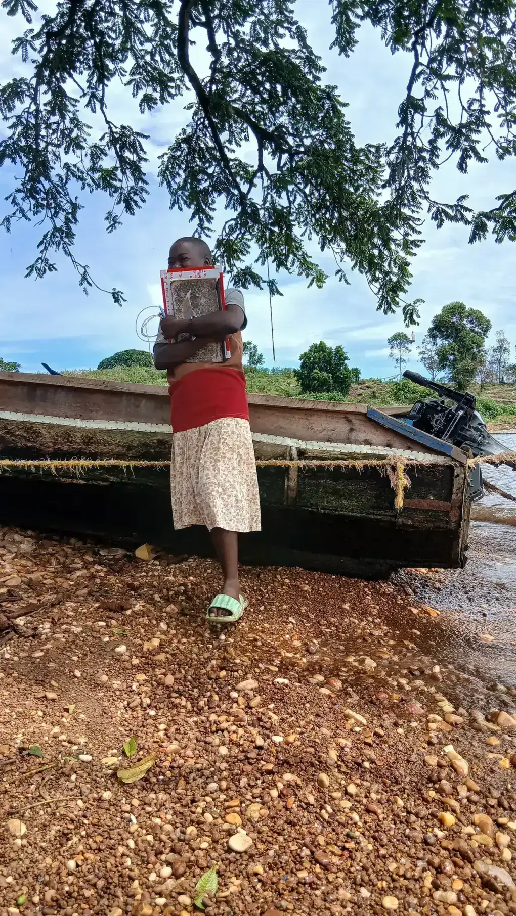 A girl in a red top holds a book to her chest while standing by a wooden fishing boat on the lakeshore