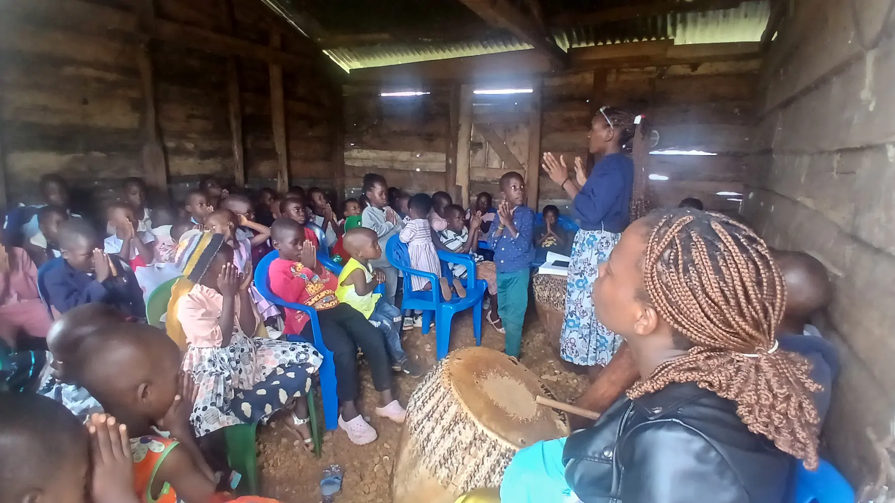 A woman leader leads a group of seated children in song inside a timber hall, a traditional drum beside her