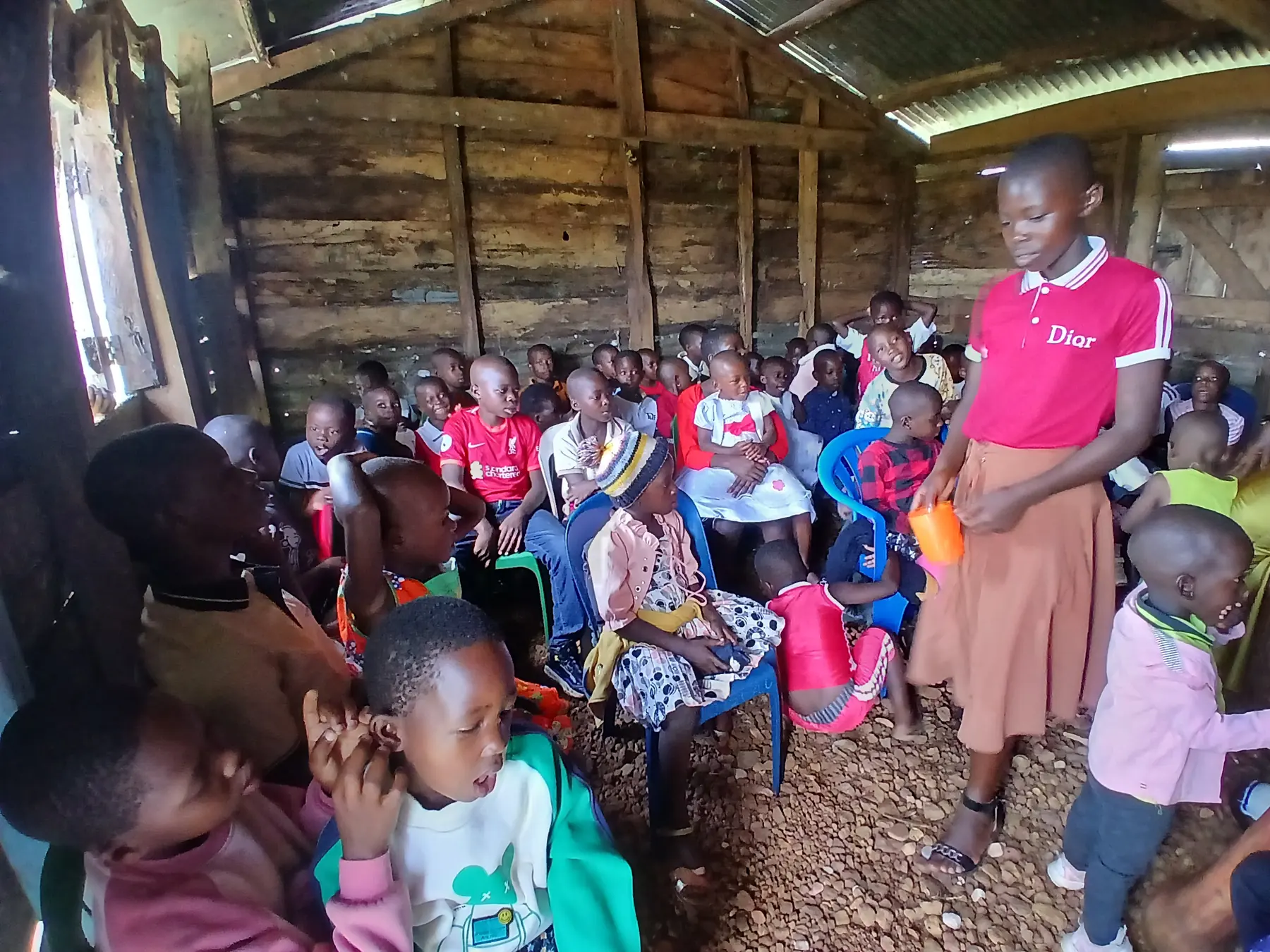 A young woman in a pink T-shirt stands among seated children inside a timber hall